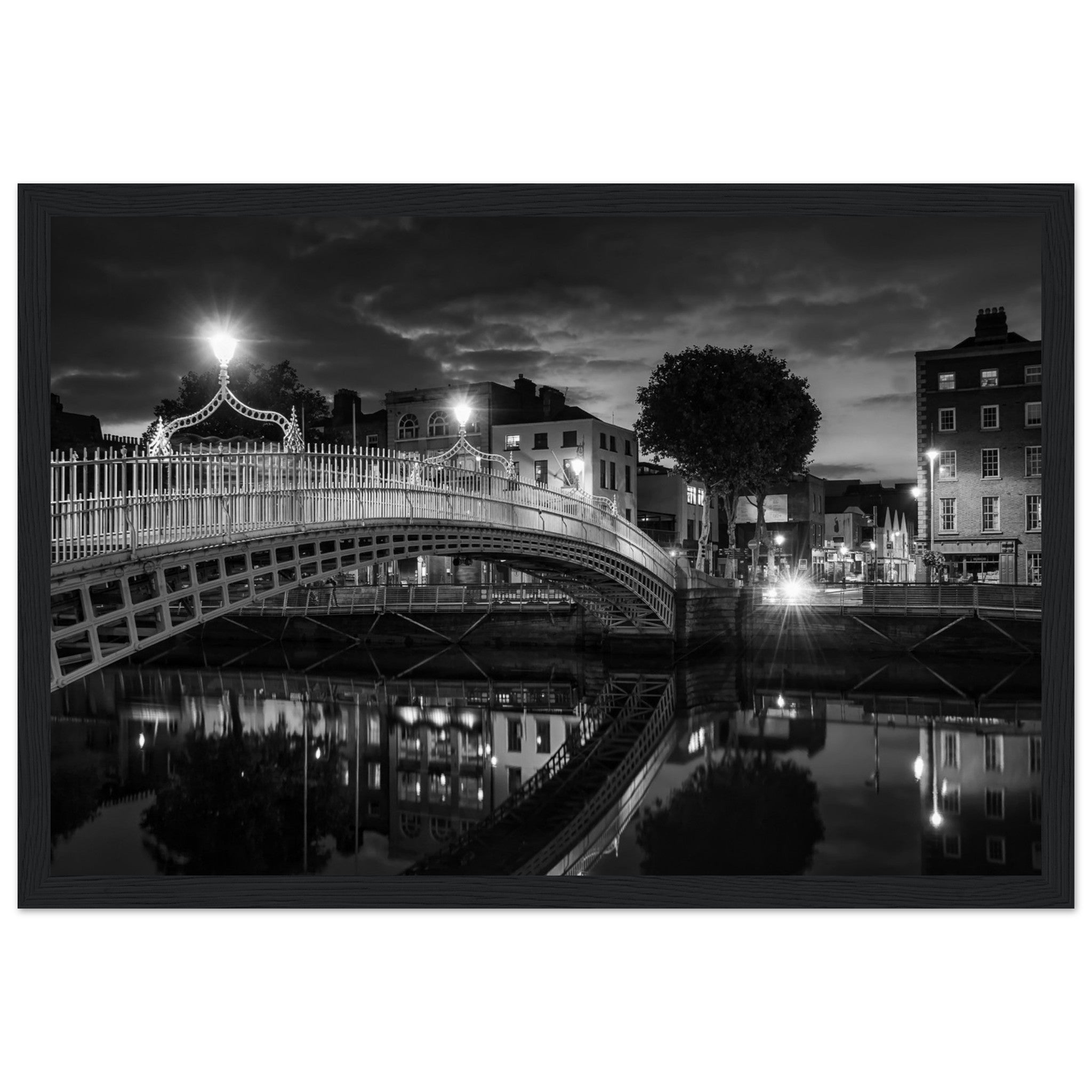  Black and white framed wall art print of the Ha'penny Bridge in Dublin at night. The bridge's historic architecture is beautifully highlighted against the dark sky, with reflections shimmering in the River Liffey below. Ideal for adding elegance to any room or office.